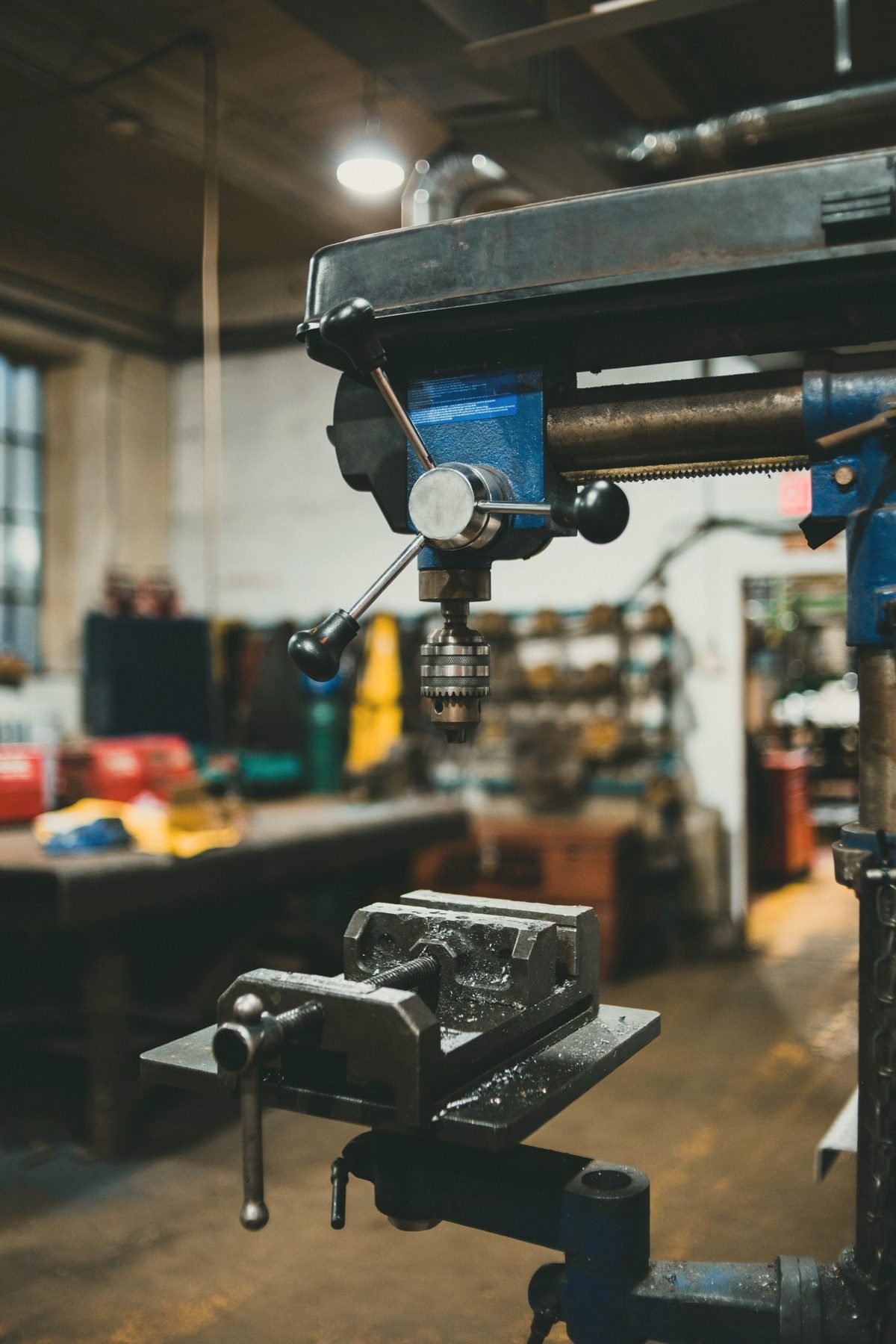 Pedestal drill press standing in a workshop with tooling on the bench behind.