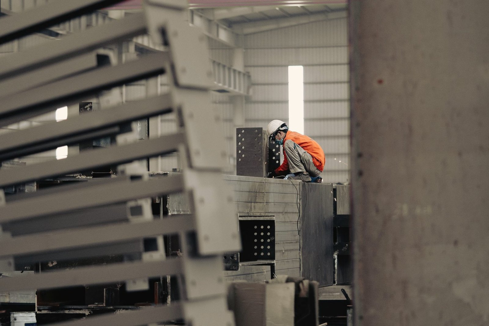 Worker in a helmet and orange vest working on heavy fabricated steel sections stacked in a shop bay.